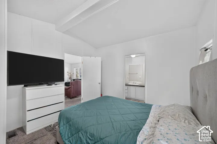 White paneled walls with a vaulted ceiling, gray carpeting, and an open doorway revealing a bathroom with a vanity and mirror