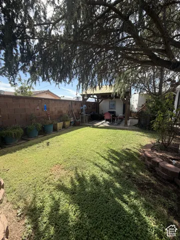 Expansive backyard featuring a manicured lawn, a mature shade tree, a covered patio area, and a block perimeter wall