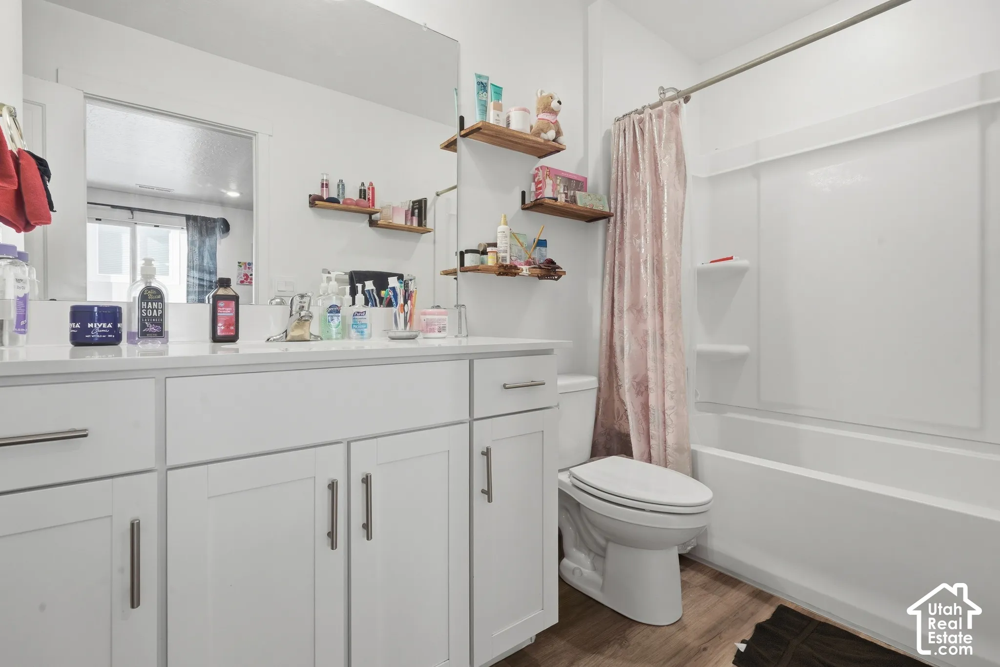 White vanity with dual sinks, brushed nickel hardware, expansive mirror, wood-finish flooring, and a white bathtub with shower