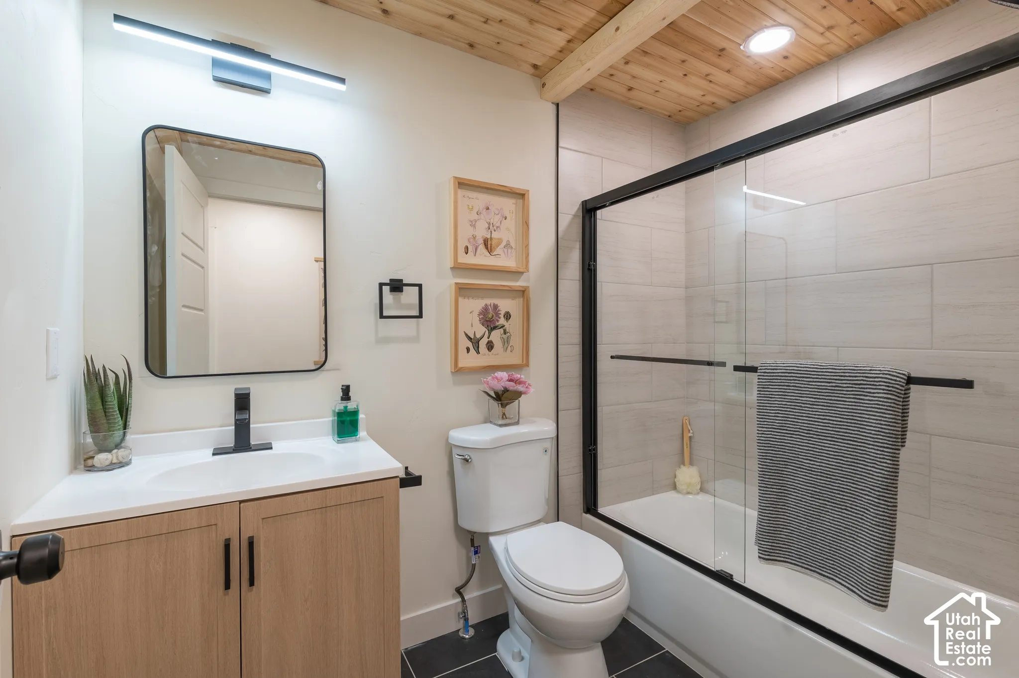 Wood-finish ceiling with exposed beams, white tile shower surround, black-framed sliding glass shower door, light wood-finish vanity with white countertop, and black-finish fixtures