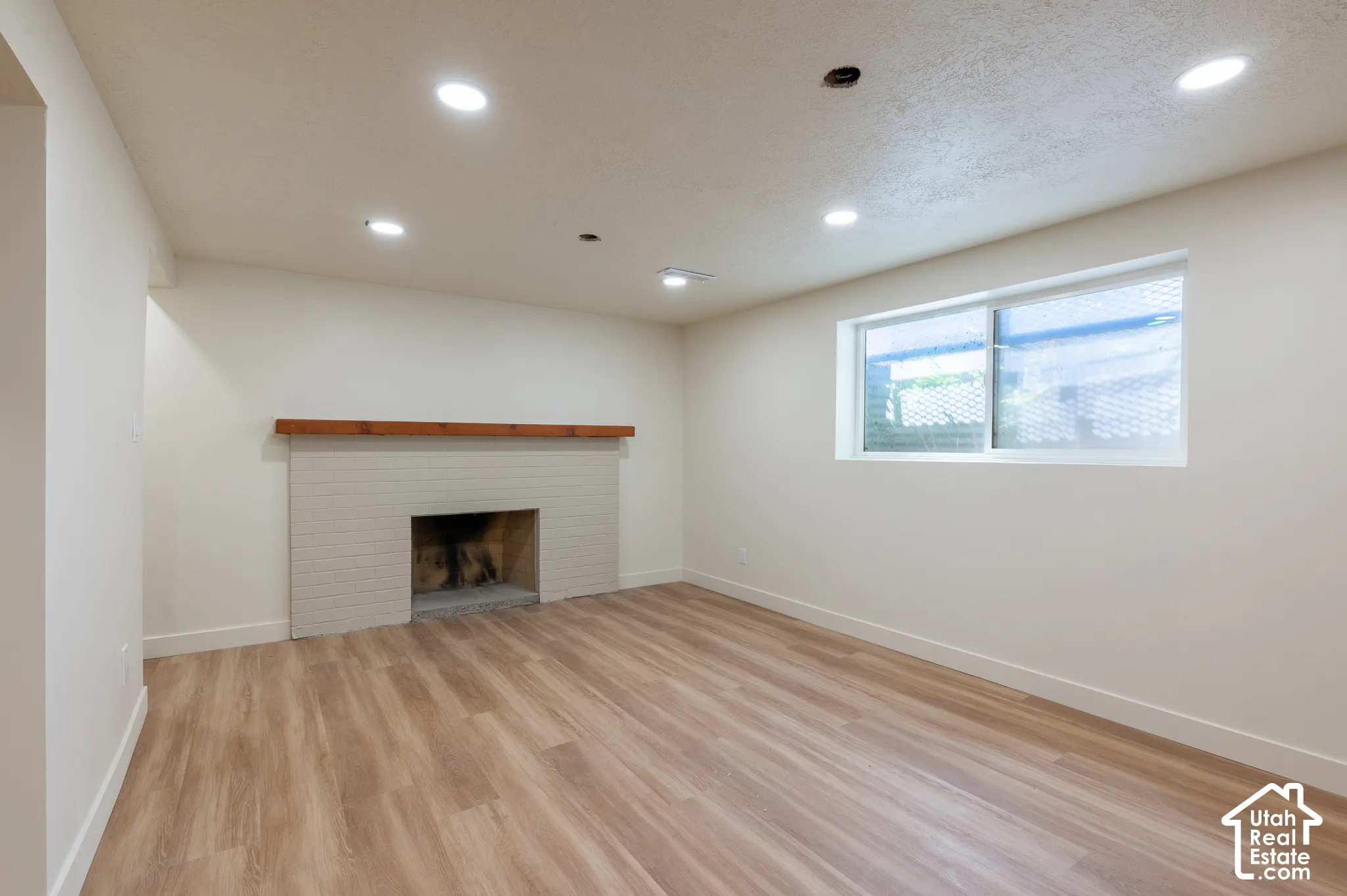 Spacious room featuring wood-finish flooring, a painted brick fireplace with a wood mantel, a window, and recessed ceiling lighting