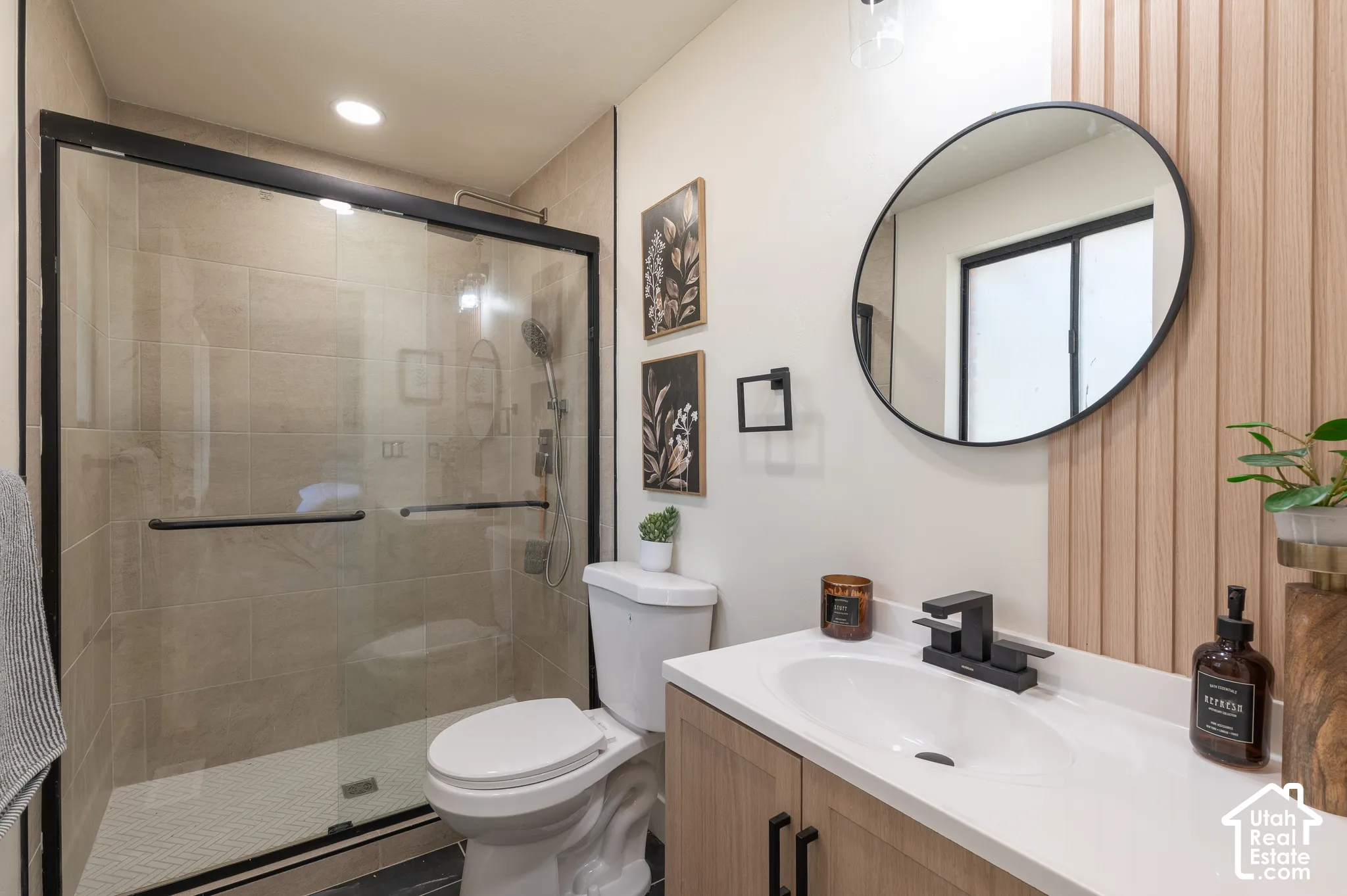 Walk-in shower with clear glass enclosure, a vanity with a white countertop and integrated sink, wood-finish cabinetry with matte black pulls, and a round black-framed mirror