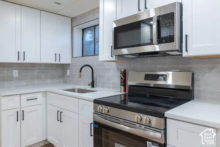 Kitchen featuring white shaker cabinetry, stainless steel appliances, light-toned countertops, a matte black and brass faucet, and a gray subway tile backsplash