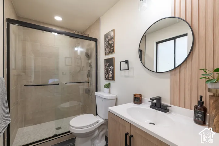 Walk-in shower with clear glass enclosure, a vanity with a white countertop and integrated sink, wood-finish cabinetry with matte black pulls, and a round black-framed mirror