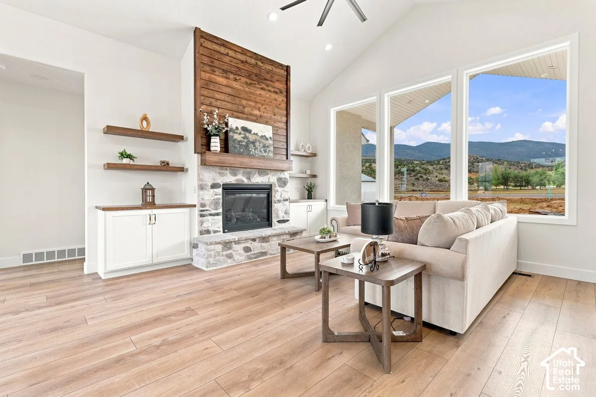 Vaulted ceiling, stone-surround fireplace with wood paneling, light wood-finish flooring, built-in cabinetry with floating shelves, and large windows