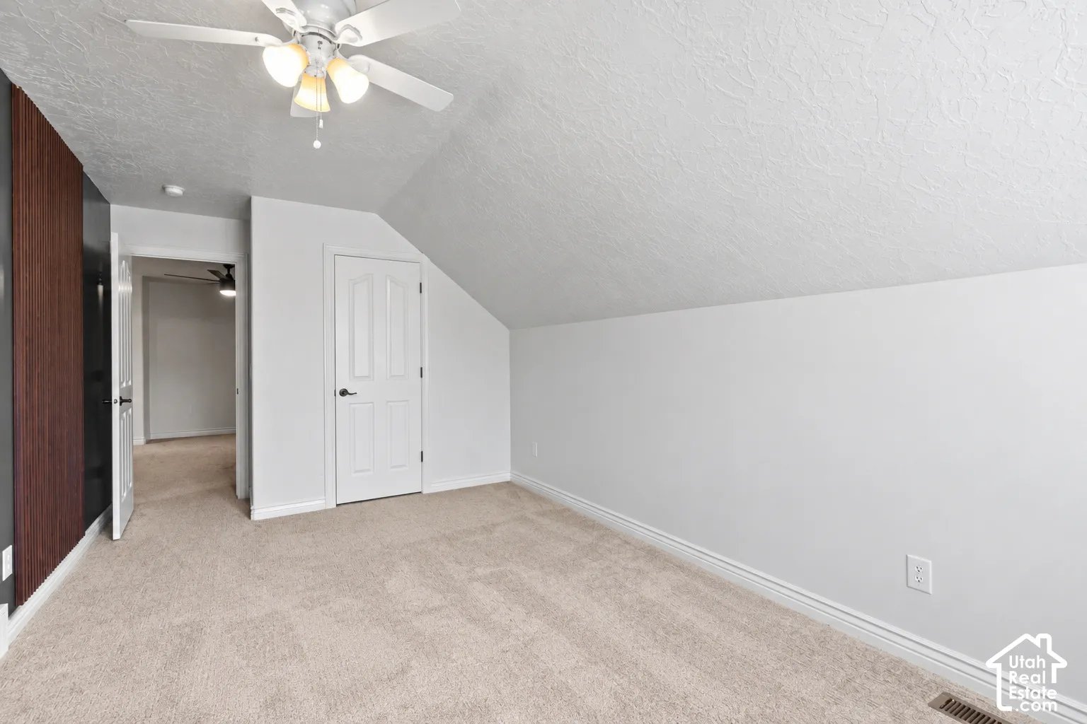 Angled ceiling with a textured finish, ceiling fan with light kit, neutral carpet flooring, white interior doors, and a wood-finish accent wall