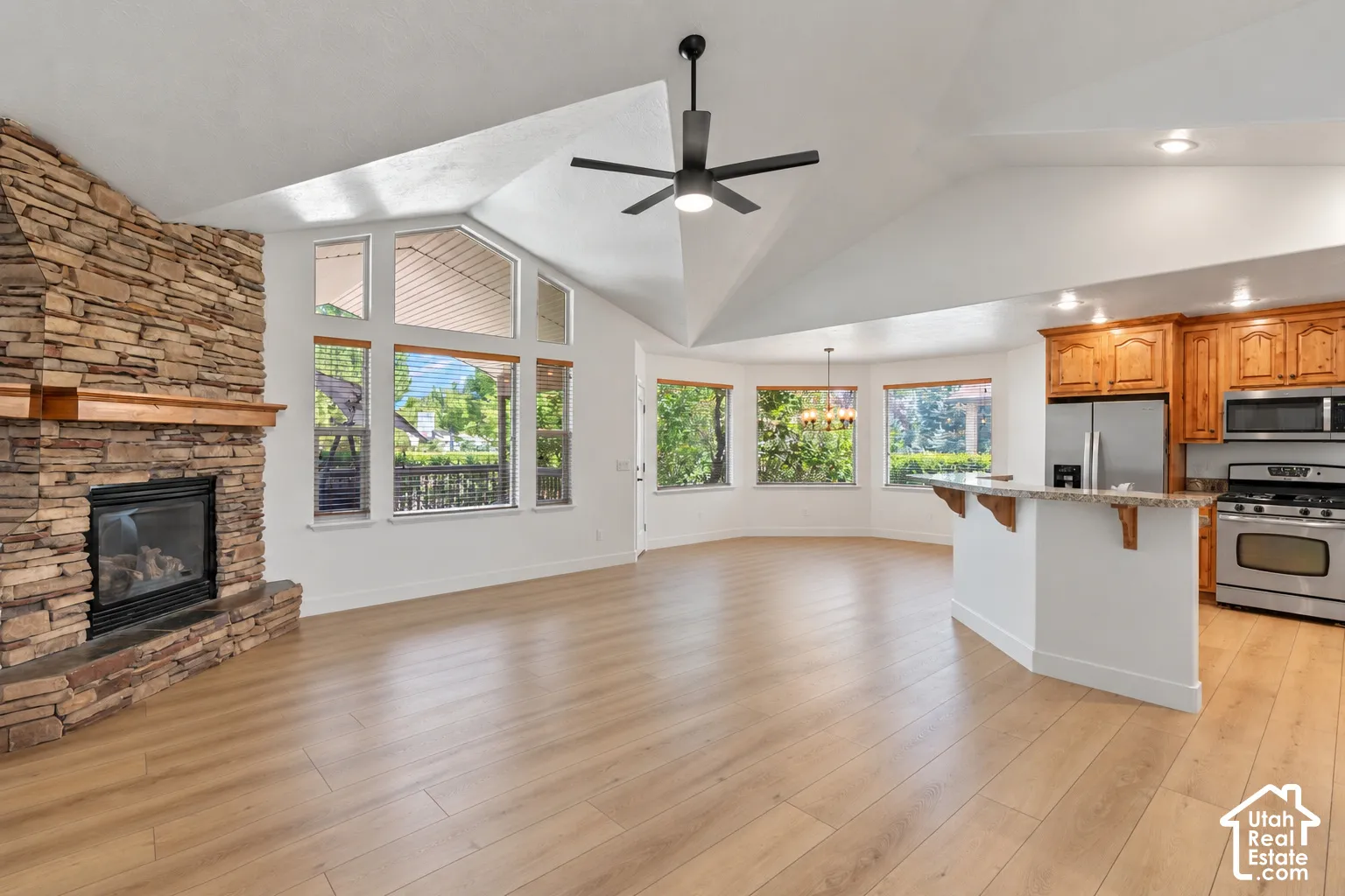 Vaulted ceilings with a ceiling fan, stone-clad fireplace with wood mantel, wood-finish flooring, and a kitchen featuring wood cabinetry and stainless steel appliances