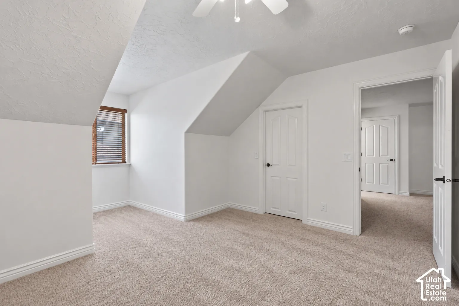 White-painted interior with sloped ceilings, textured walls, light-toned carpeting, and a window with wood blinds