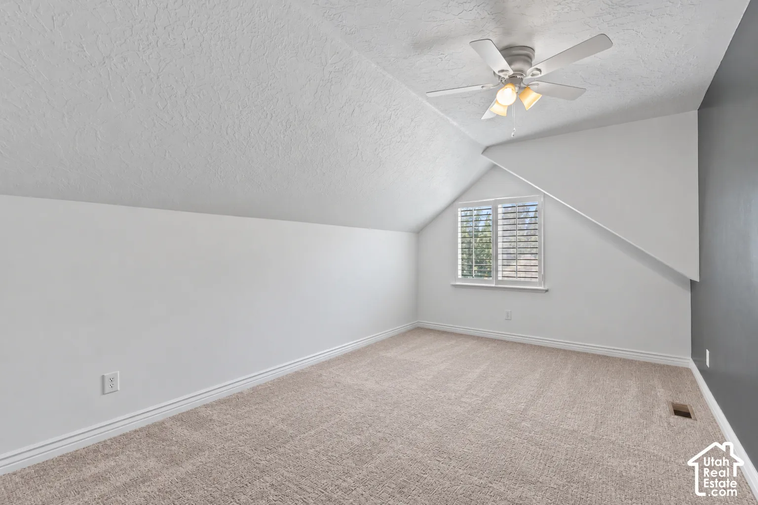 Angled ceiling room with neutral carpeting, a ceiling fan, a window featuring white plantation shutters, and a contrasting accent wall