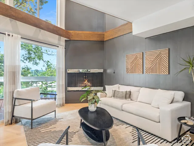 Vaulted ceilings with exposed wood beams, expansive windows, light wood-finish flooring, and a built-in fireplace with a metal screen