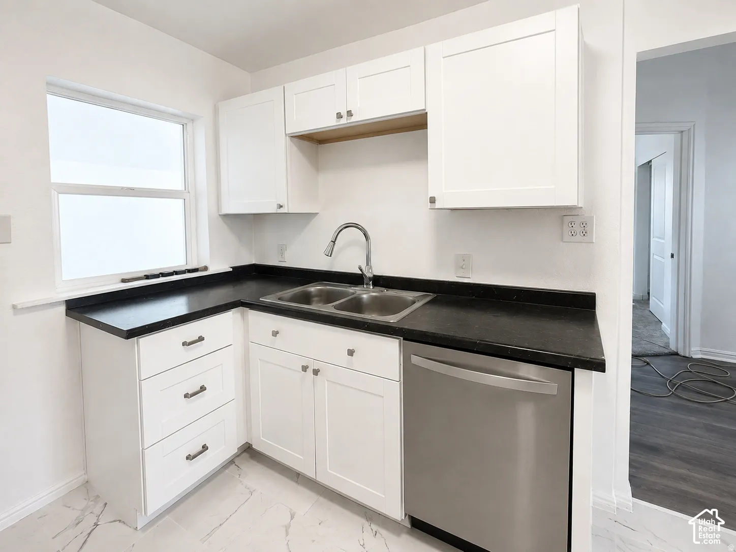 Kitchen featuring white shaker-style cabinetry, dark countertops, a double basin sink with a gooseneck faucet, and a stainless steel dishwasher