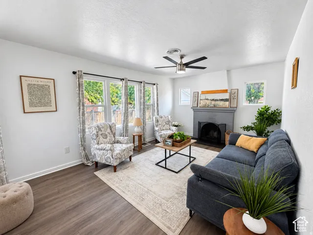 Wood-finish flooring, multiple windows, centered fireplace, ceiling fan, and white walls