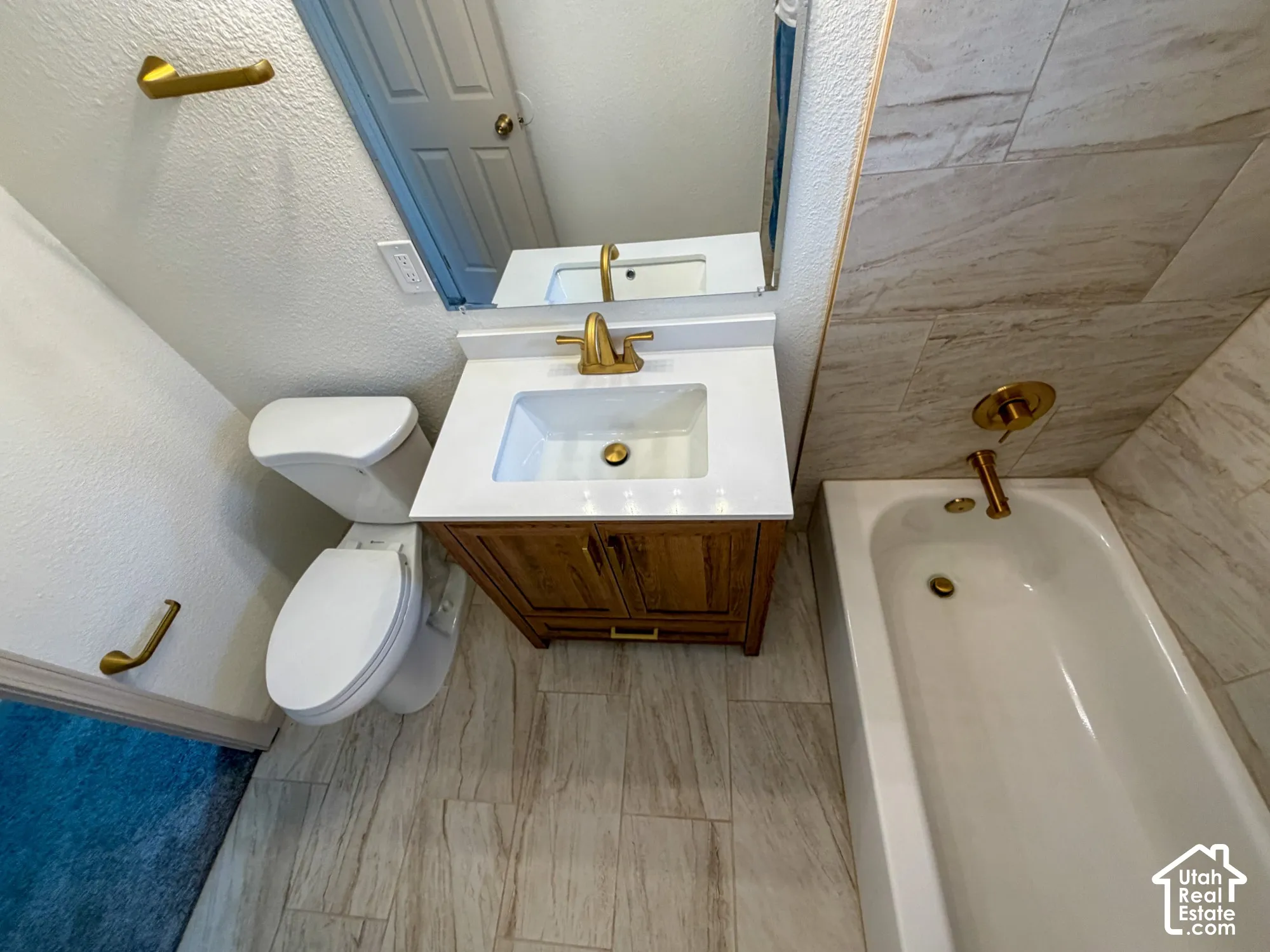 Wood-finish tile flooring, white vanity with gold-tone faucet, white bathtub with gold-tone fixtures, and tile wall surround
