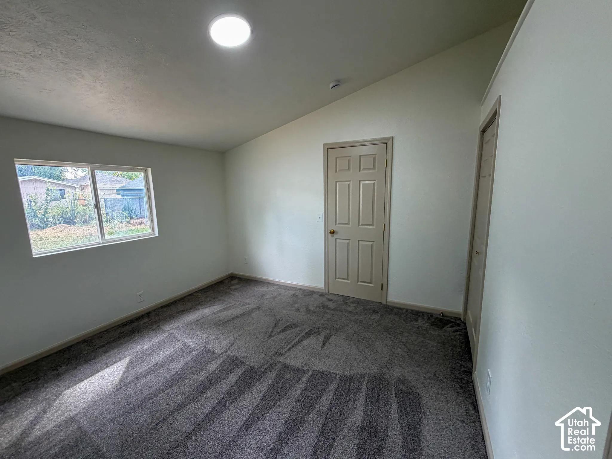 Vaulted ceiling with an overhead light fixture, light-colored walls, window with outdoor scenery, and patterned carpet flooring