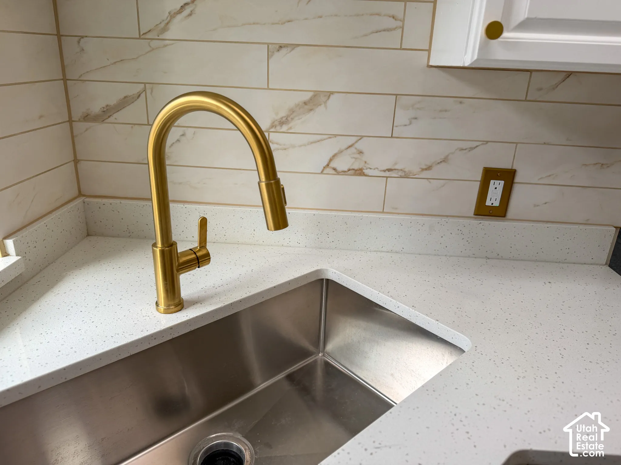 Stainless steel sink, white speckled countertop, gold-finish faucet, and tiled backsplash