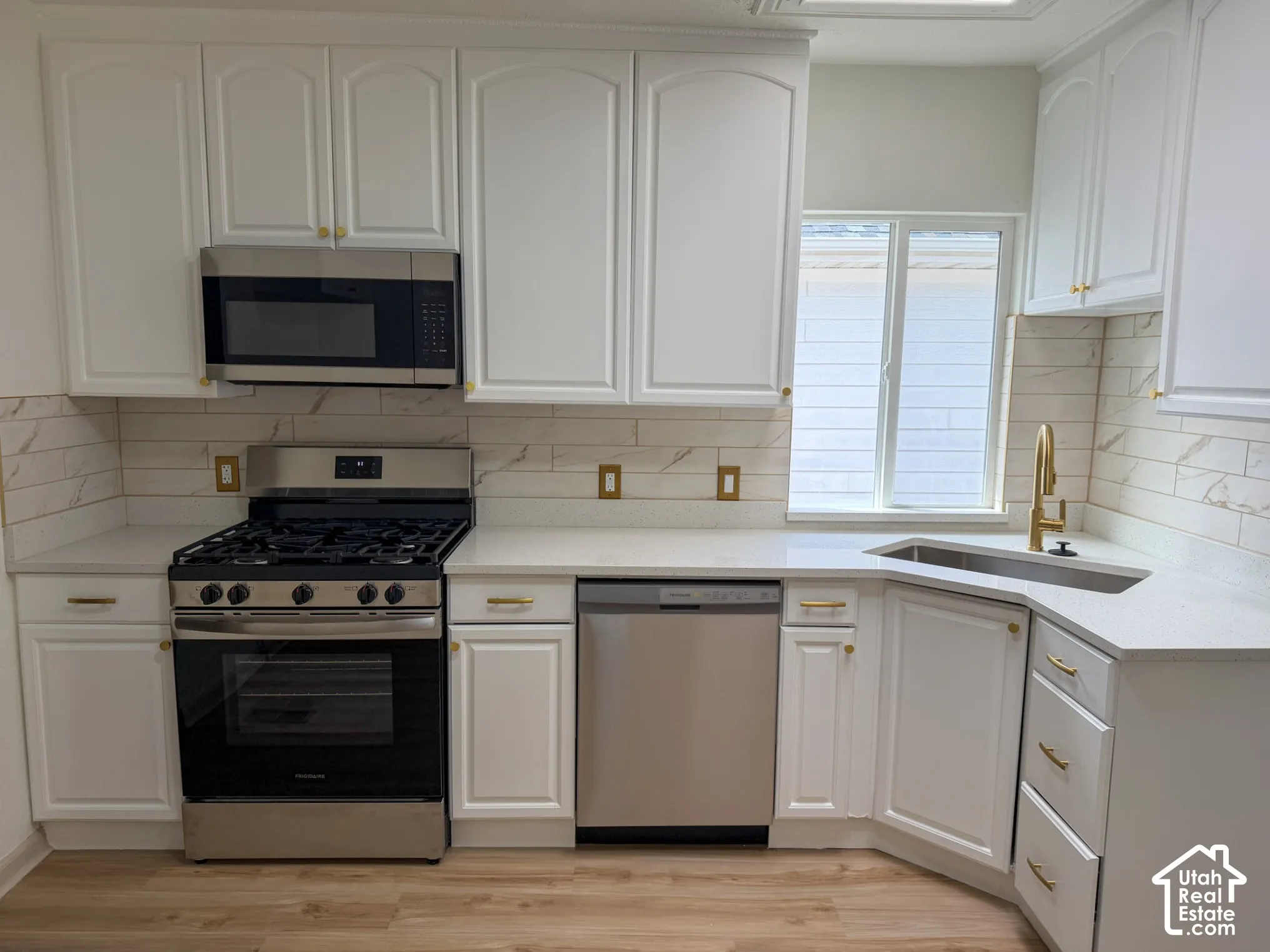 Kitchen featuring white cabinetry with gold-finish hardware, stainless steel appliances, a white tile backsplash, light countertops, and wood-finish flooring