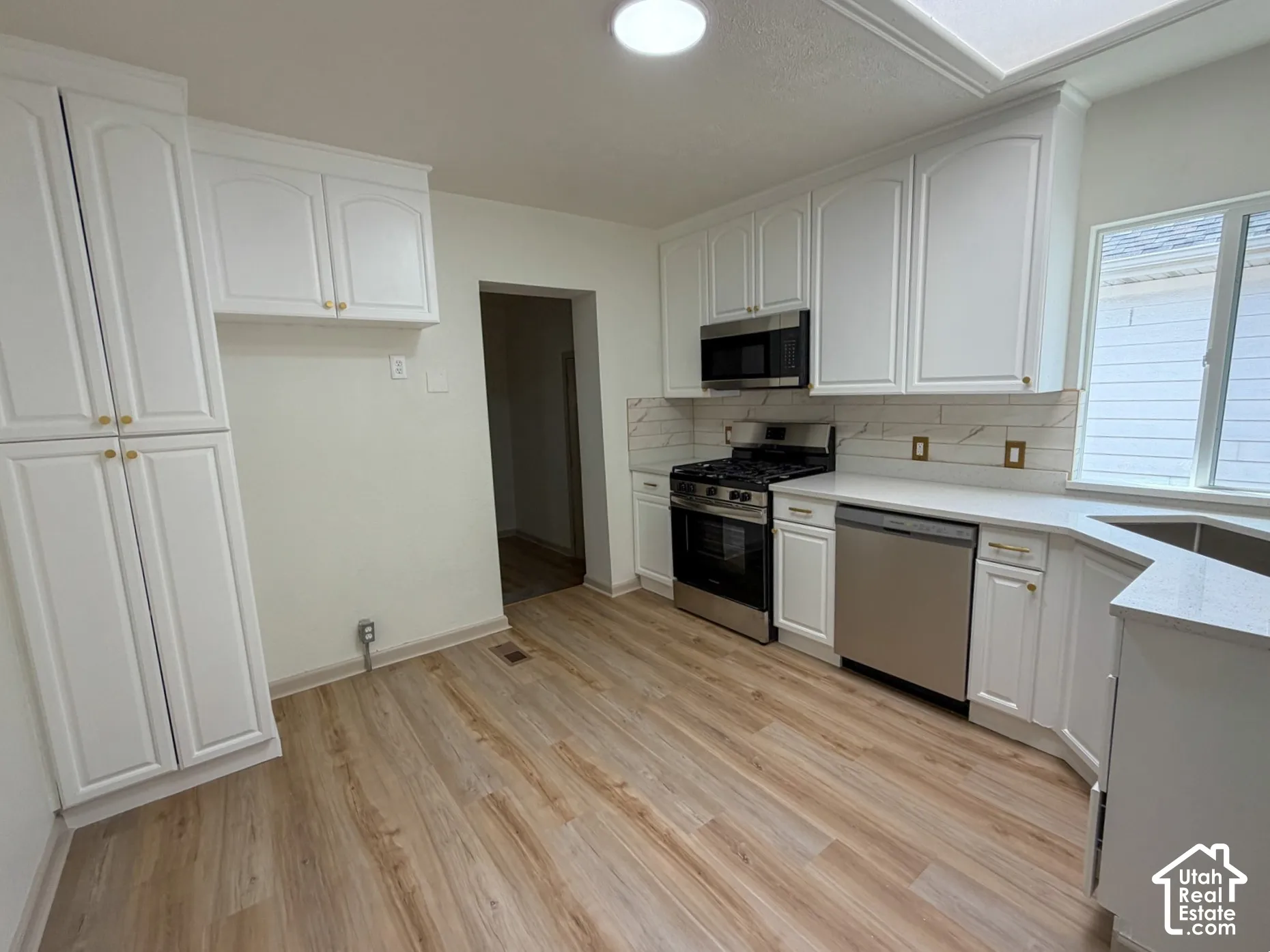 Kitchen featuring wood-finish flooring, white cabinetry with gold-tone hardware, stainless steel appliances, white subway tile backsplash, and light-toned countertops