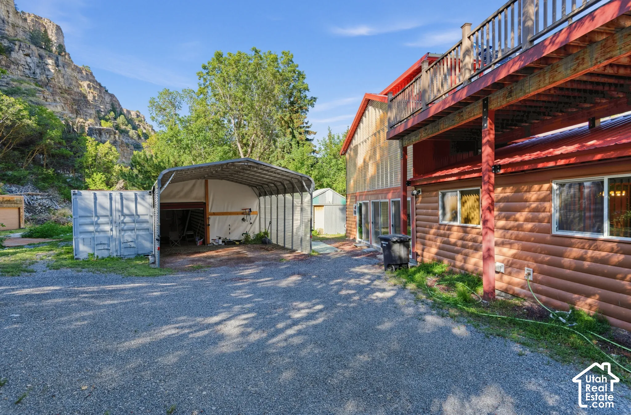 Gravel driveway, log-style exterior siding, red metal roof, upper-level deck with wood railing, covered carport, and a container storage unit