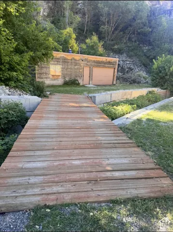 Wood-plank bridge with natural surroundings, leading to a structure with a roll-up door and a window