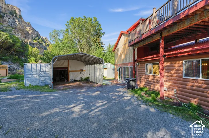 Gravel driveway, log-style exterior siding, red metal roof, upper-level deck with wood railing, covered carport, and a container storage unit