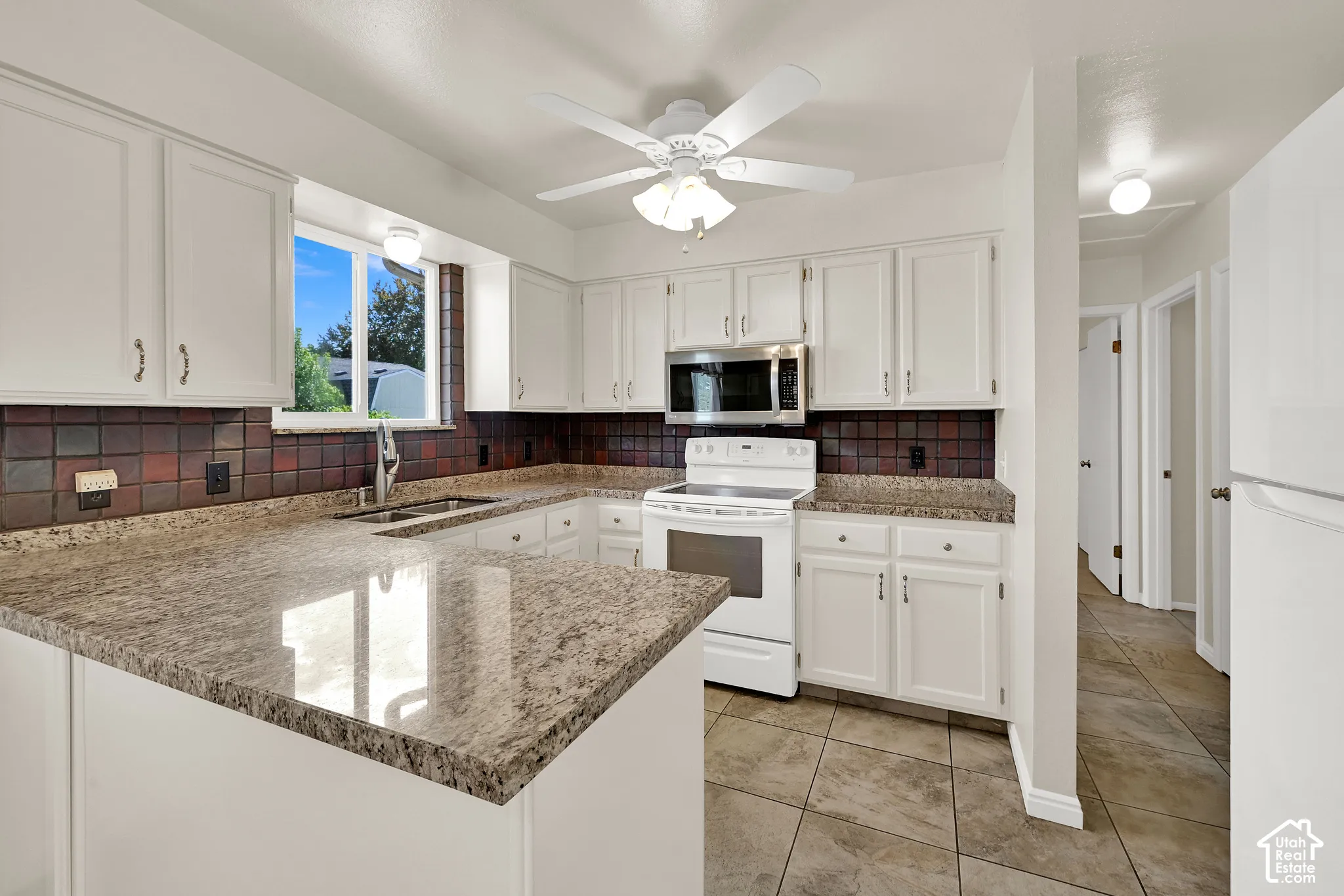 Bright, open kitchen featuring white cabinetry with brushed nickel hardware, granite countertops, a large island with seating space, stainless steel appliances, a tiled backsplash and a ceiling fan