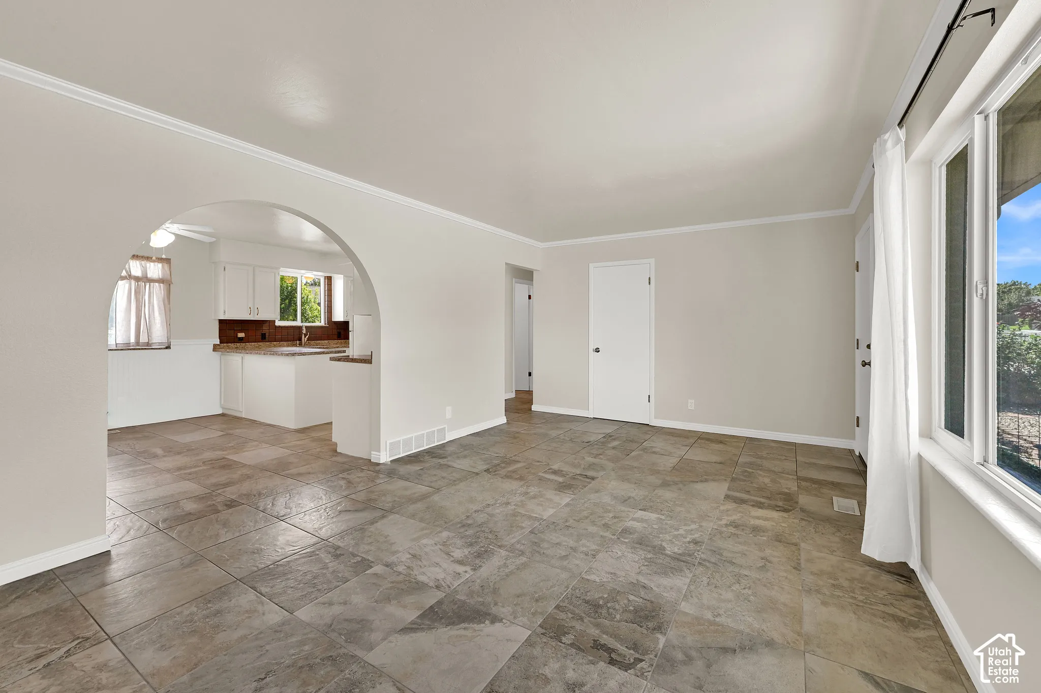 Spacious open-concept living area showcasing tile flooring, abundant natural light from multiple windows, arched architectural detailing, and seamless sightlines to the kitchen with built-in cabinetry and countertop workspace
