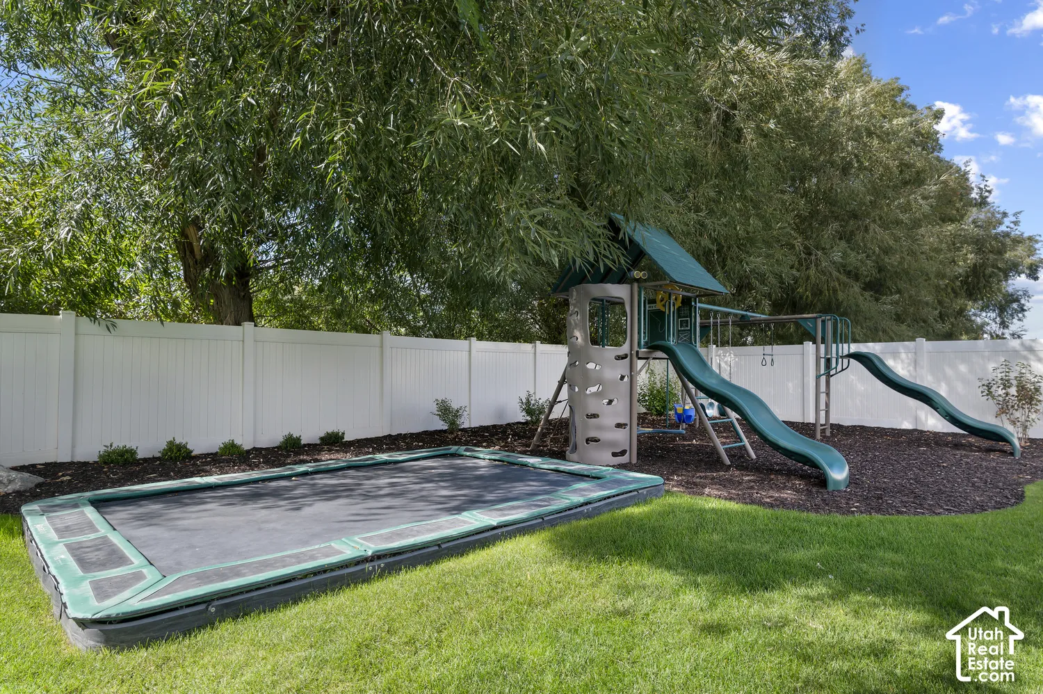 Expansive yard featuring a manicured lawn, white privacy fencing, mature trees, and a built-in trampoline