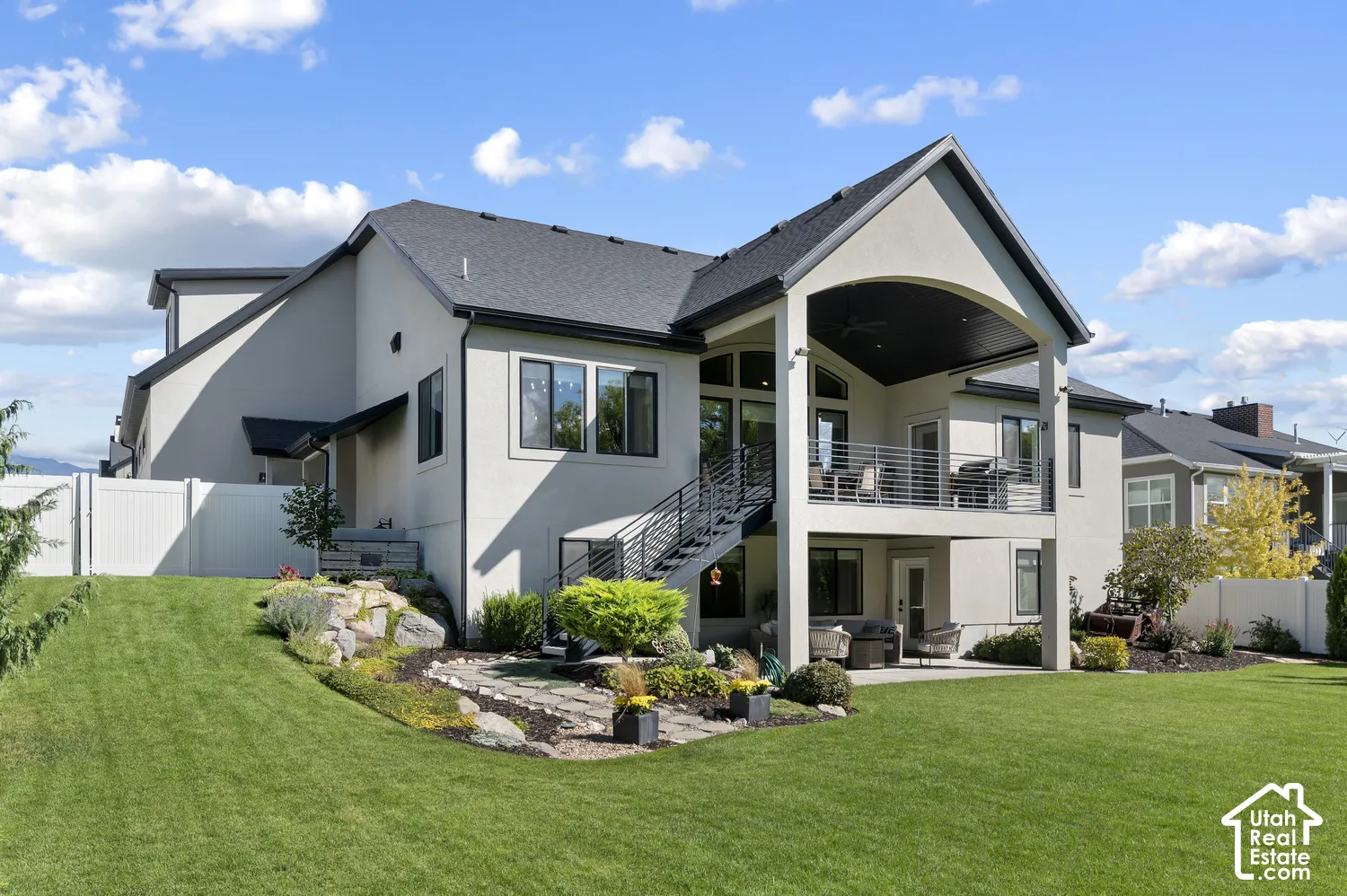 Expansive backyard with a manicured lawn, multi-level stucco exterior, upper-level covered patio with metal railings, and a lower-level patio