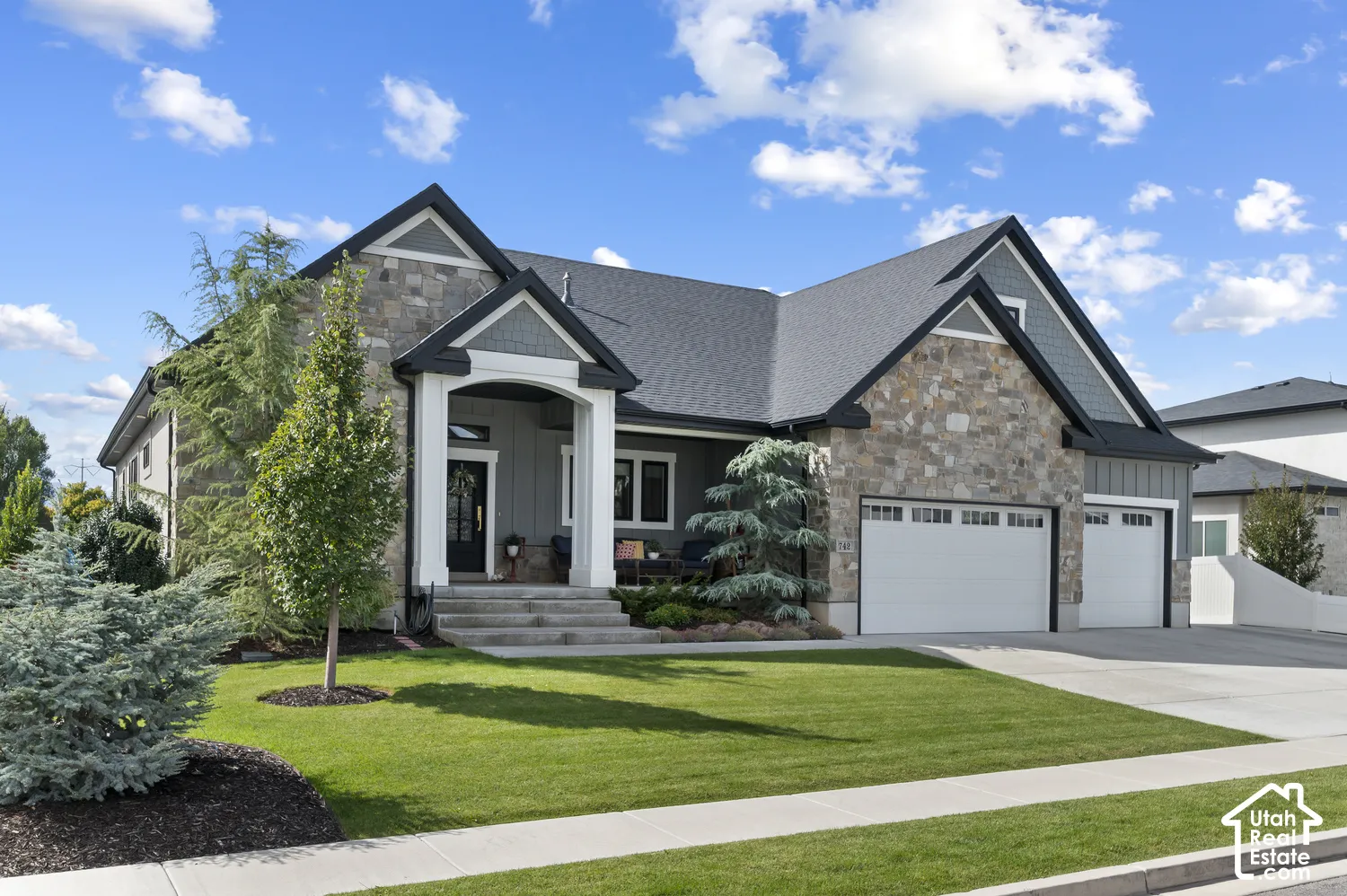 Stone facade, covered entry, attached multi-car garage, concrete driveway, and manicured landscaping