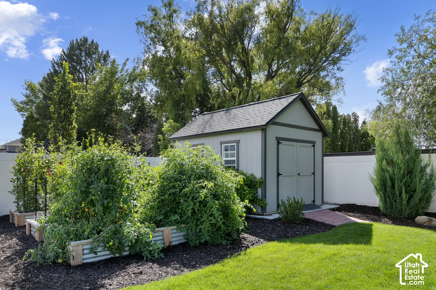 Manicured lawn with raised garden beds, a storage shed with a shingled roof, a brick pathway, and mature landscaping