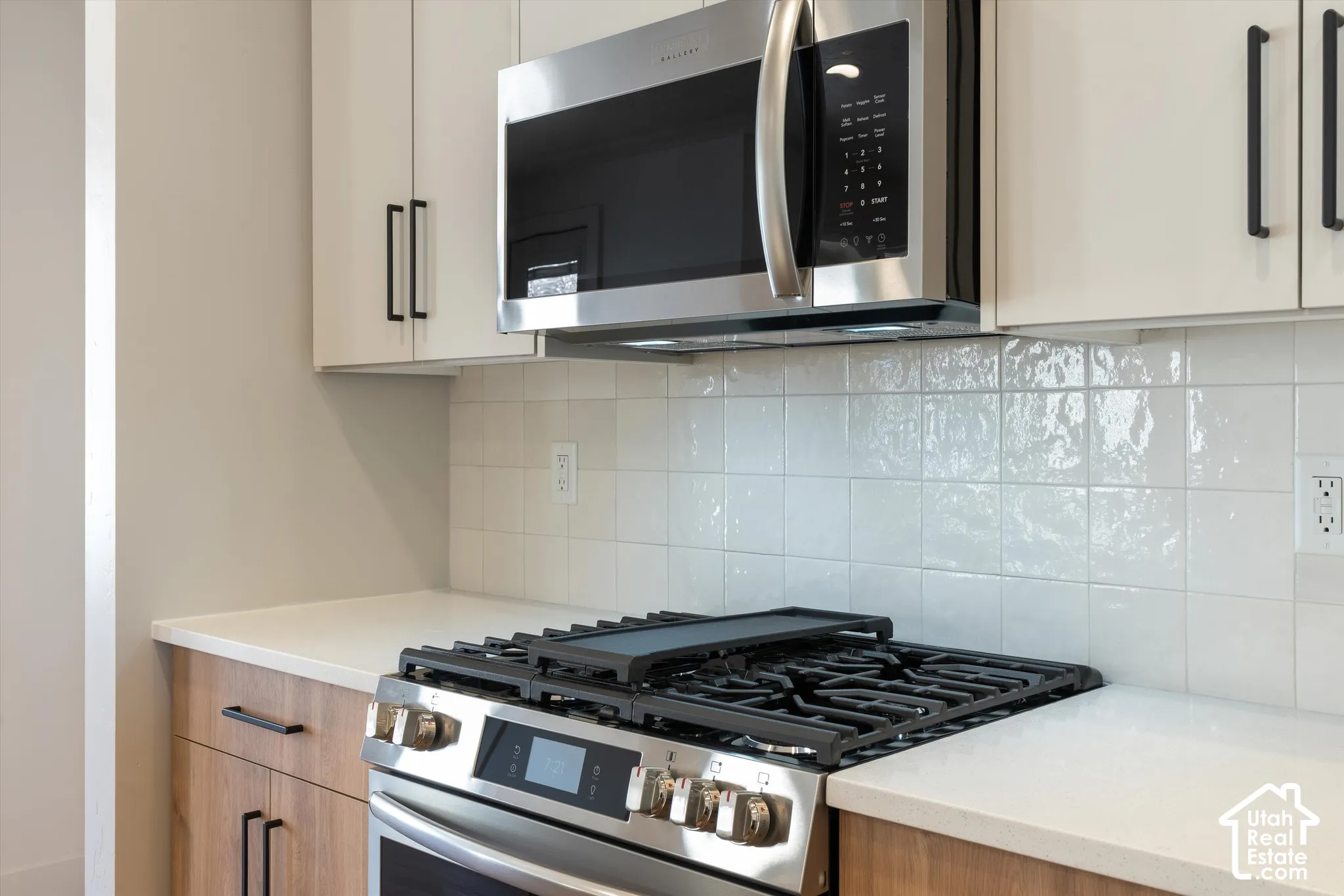 Stainless steel microwave, gas range, wood-finish lower cabinetry, white upper cabinetry, and white tile backsplash