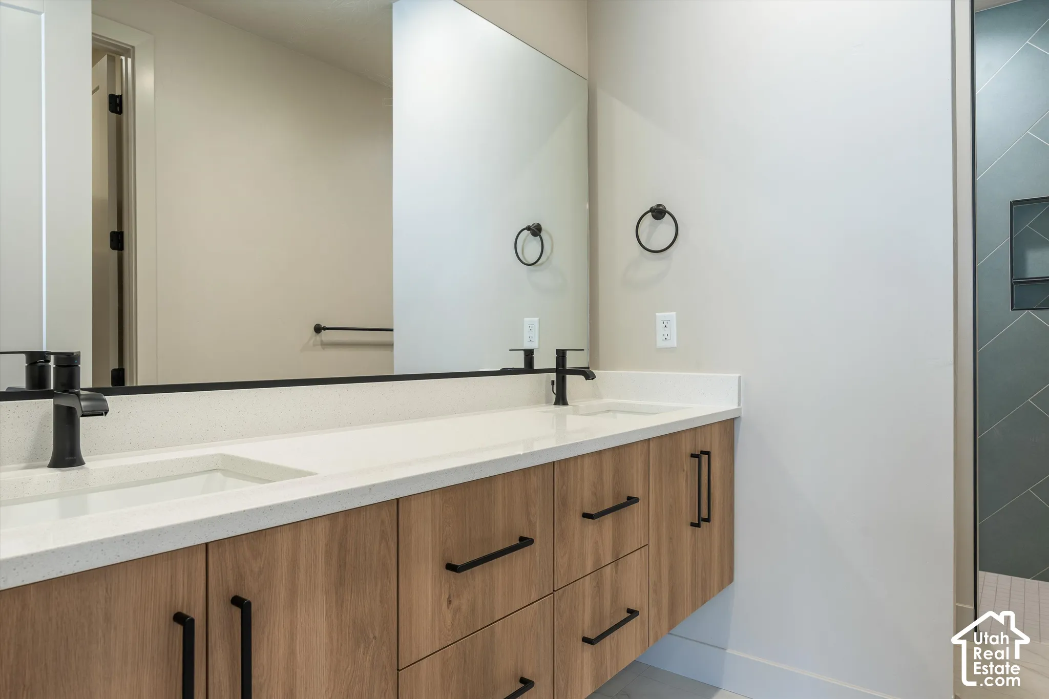 Floating wood-finish vanity with dual integrated sinks, matte black fixtures, full-width mirror, and a walk-in shower featuring patterned tile