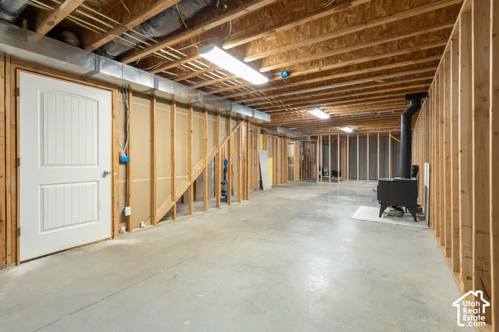 Unfinished basement featuring exposed wood framing, concrete flooring, overhead fluorescent lighting, and a freestanding wood-burning stove