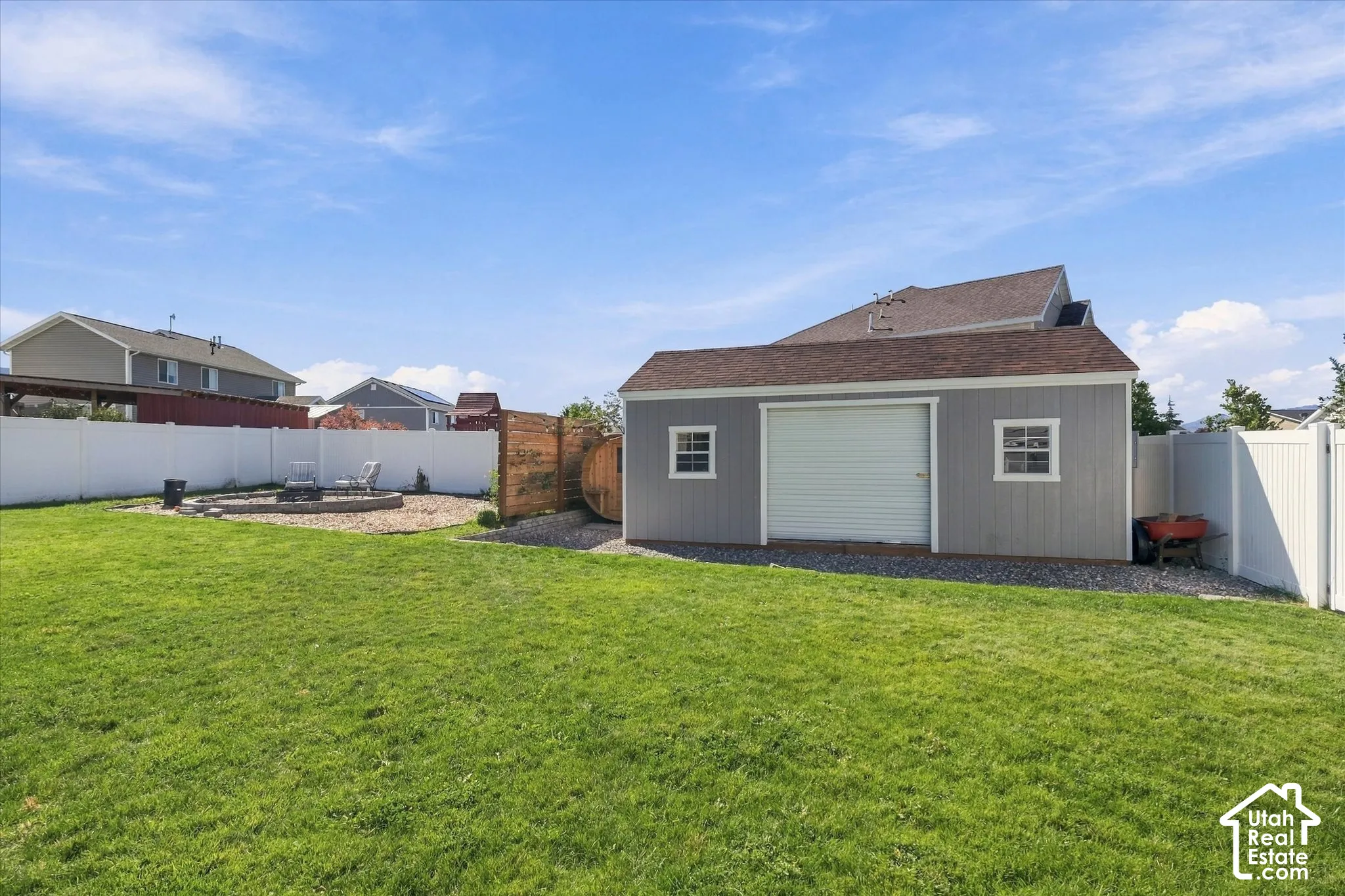 Expansive lawn with a white vinyl privacy fence, a detached storage shed featuring a roll-up door, and a circular gravel patio with a built-in fire pit