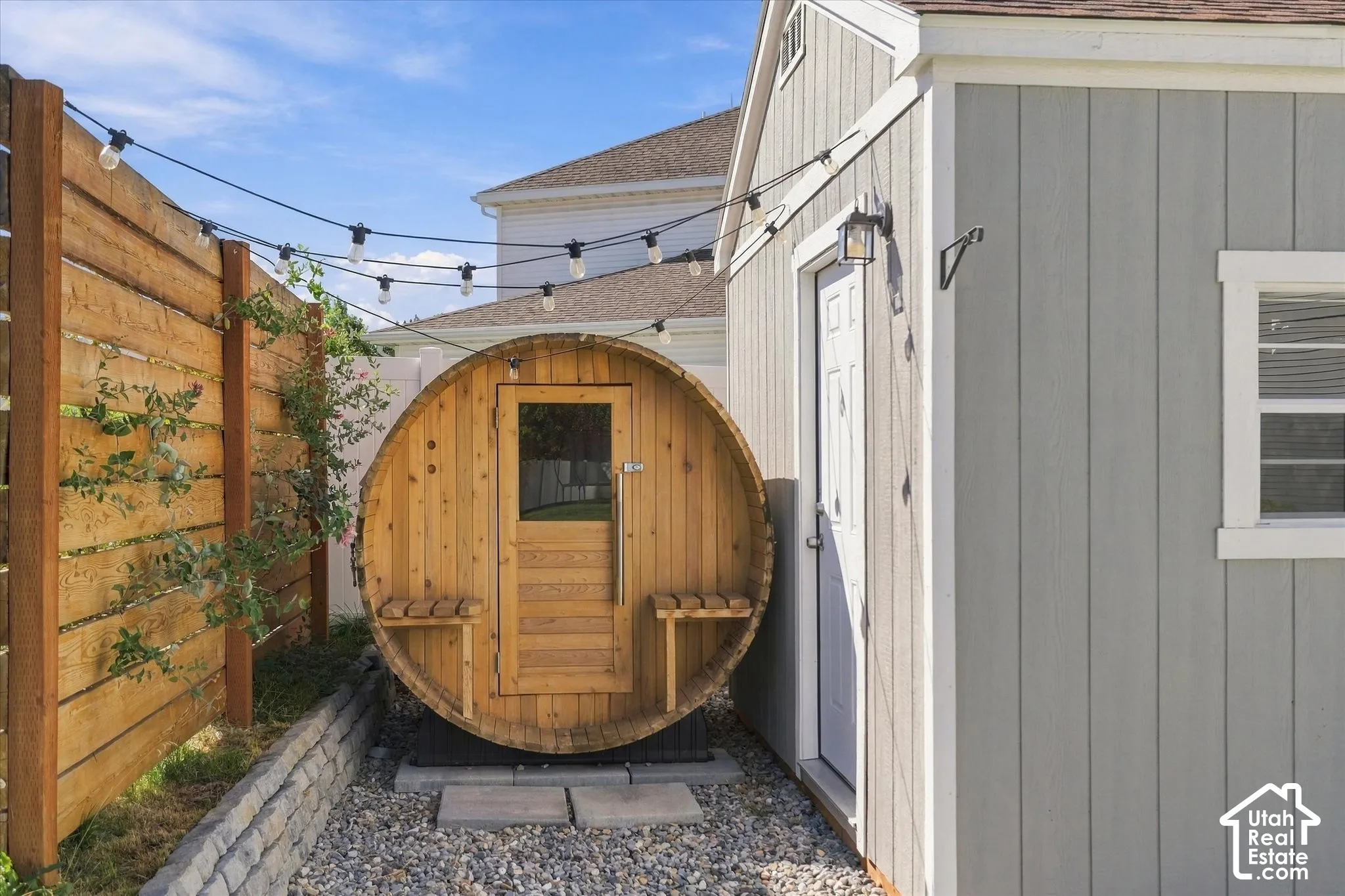 Barrel-style sauna with exterior seating, wood privacy fence, gravel ground cover, and string lighting