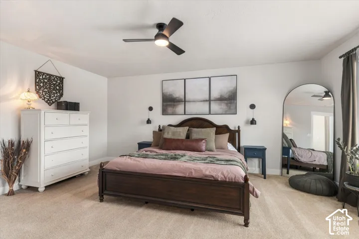 Plush carpet flooring, ceiling fan with integrated lighting, dark wood bed frame, white walls, and dark window treatments