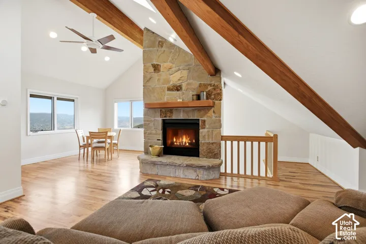 Vaulted ceiling with exposed wood beams, stone-clad fireplace with wood mantel, wood-finish flooring, and multiple windows