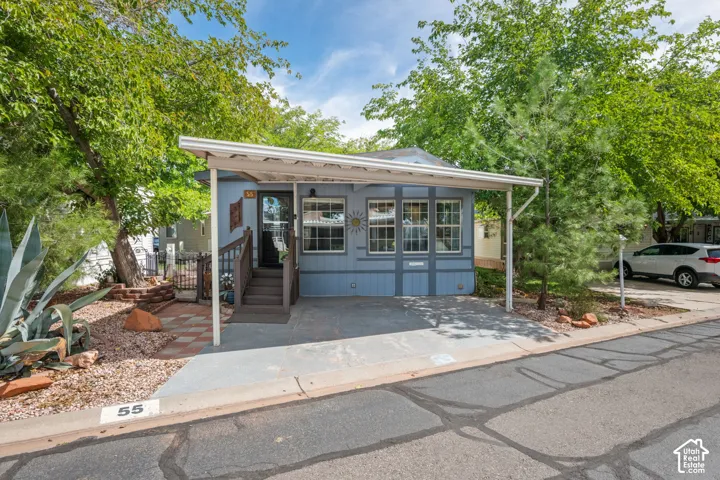 Covered carport with paved surface, front entry with wood steps and railing, multiple windows on the facade, exterior landscaping with desert plantings, and mature trees