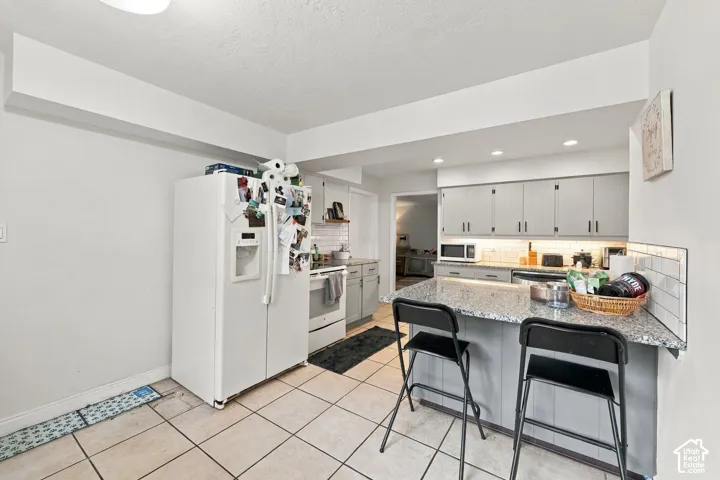 Kitchen featuring tile flooring, a built-in breakfast bar with a speckled countertop, recessed lighting, white appliances, and light gray cabinetry