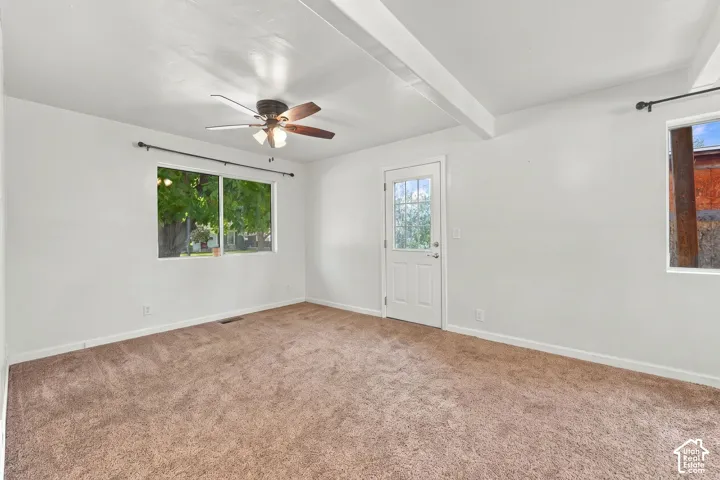 Room featuring a wood-finish ceiling fan, broad windows, exposed ceiling beams, and light-colored carpeting