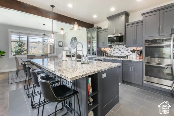 Kitchen featuring gray cabinetry, stainless steel appliances, a multi-tone tile backsplash, and a large island with a light-colored countertop