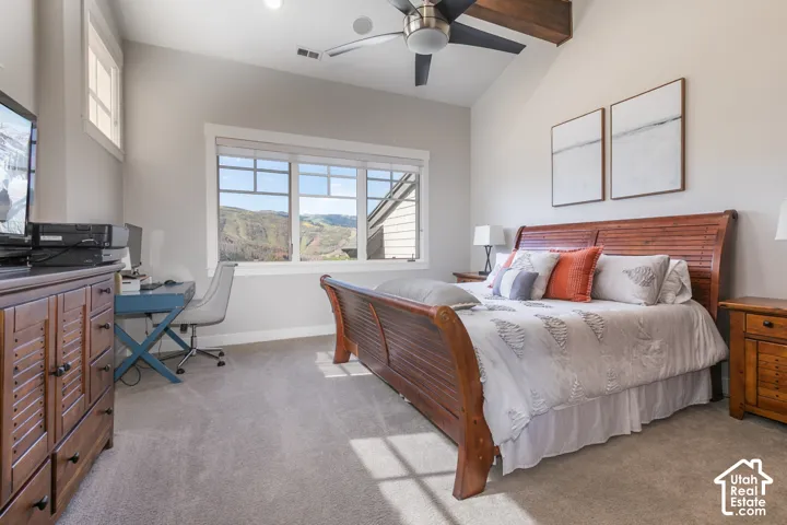 Vaulted ceiling with exposed wood beam, ceiling fan, large multi-pane window, and high-pile carpet flooring