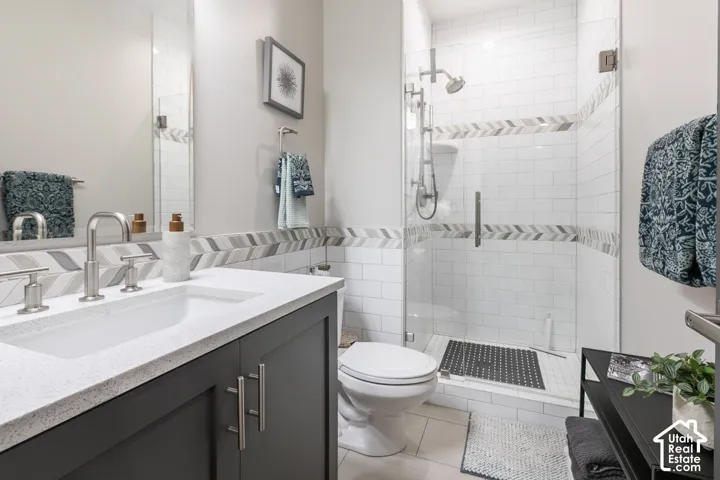 Vanity with white countertop and integrated sink, brushed nickel widespread faucet, dark gray cabinet with brushed nickel pulls, tiled shower with clear glass enclosure and chevron accent, and light-toned floor tile