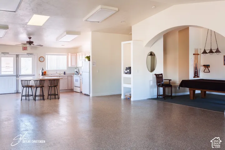 Spacious entertainment area featuring a full kitchen with white cabinetry, a built-in kitchen island, a ceiling fan, and an arched entryway leading to a dedicated game area