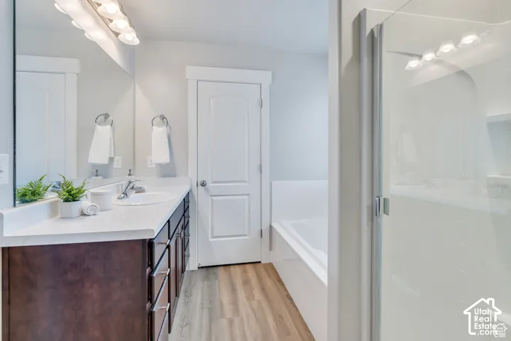Bathroom featuring a vanity with an integrated sink, a built-in bathtub, a glass-enclosed shower, wood-finish flooring, and a large mirror