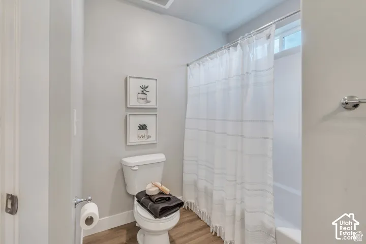 White toilet, chrome toilet paper holder, wood-finish flooring, shower/tub combination, and a high-set window