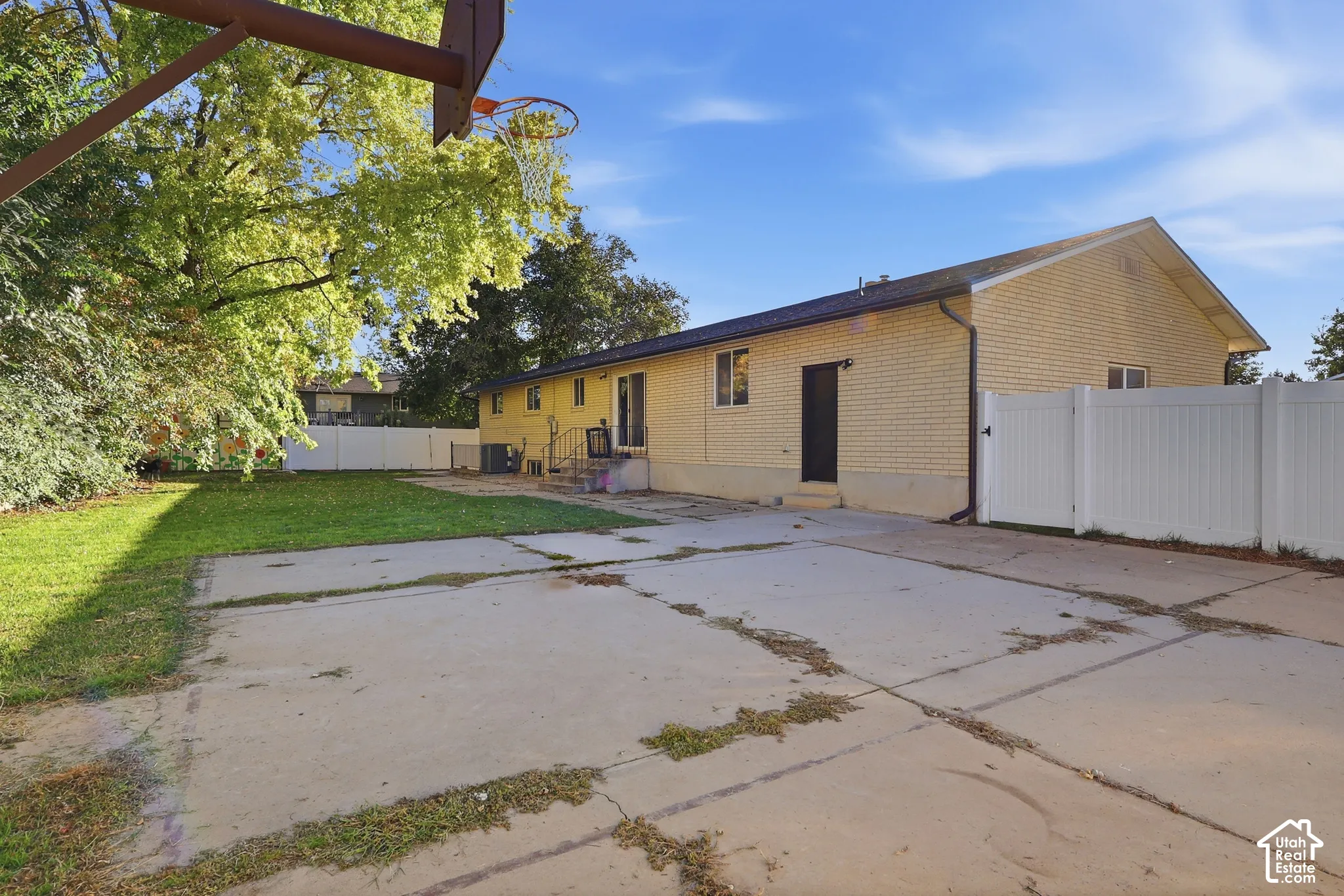 Expansive concrete patio with a perimeter white vinyl fence, an attached basketball hoop, a manicured lawn, and mature trees