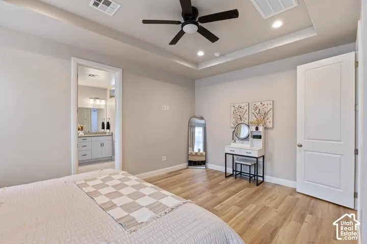 Spacious primary bedroom featuring hardwood flooring, a tray ceiling with recessed lighting and ceiling fan, and an ensuite bathroom with dual vanity, framed mirror, and tile flooring