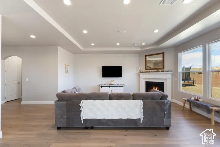Recessed lighting throughout, wood-finish flooring, corner fireplace with white mantel, tray ceiling detail, and arched doorway