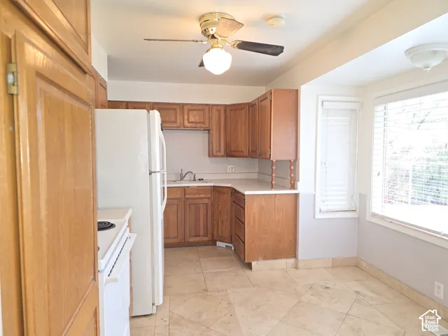 Kitchen featuring wood cabinetry, solid-surface countertops, stone-finish tile flooring, a dual-pane bay window, and a ceiling-mounted light fixture with integrated fan